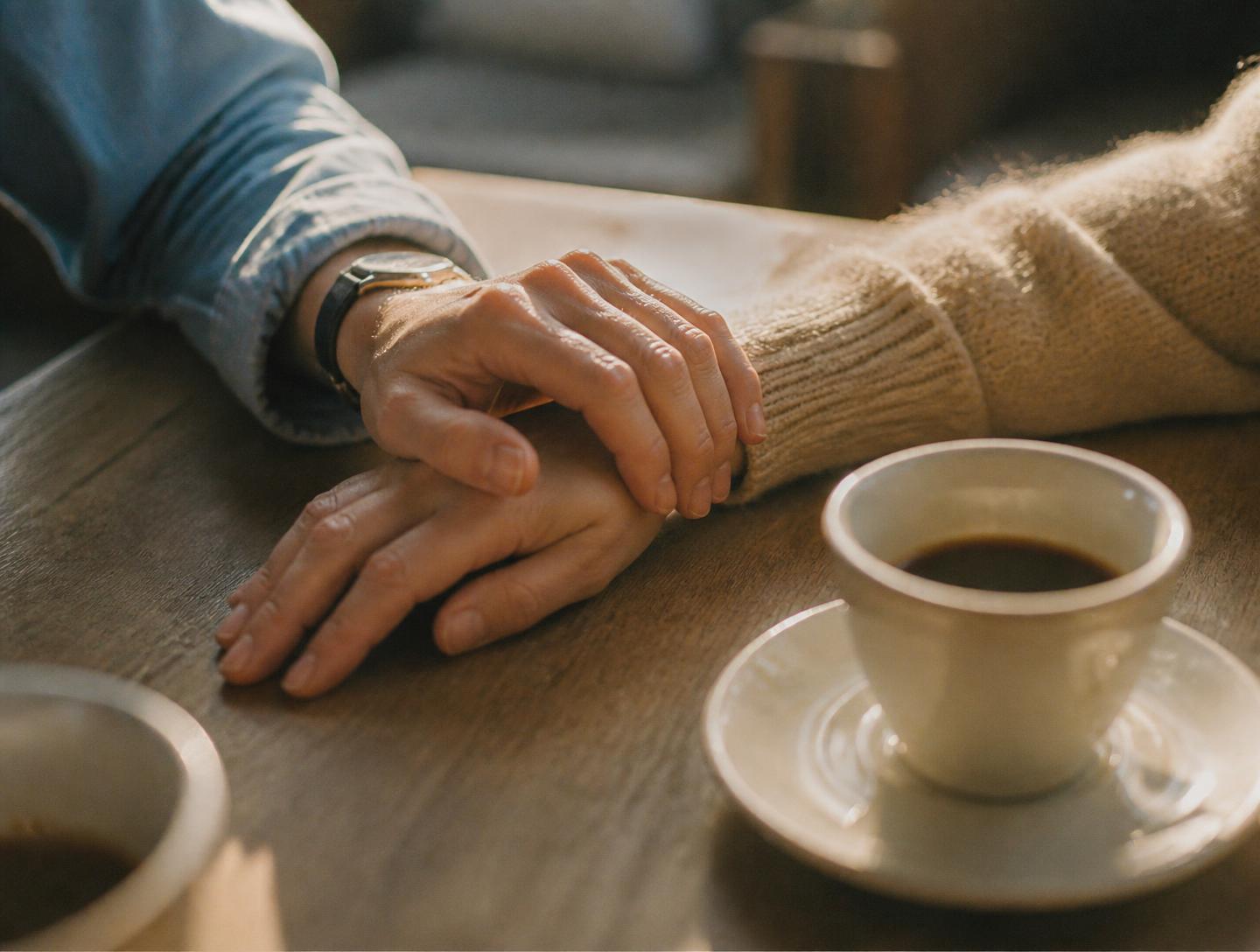 Dos manos apoyadas juntas sobre una mesa de madera, junto a una taza de café, en luz cálida